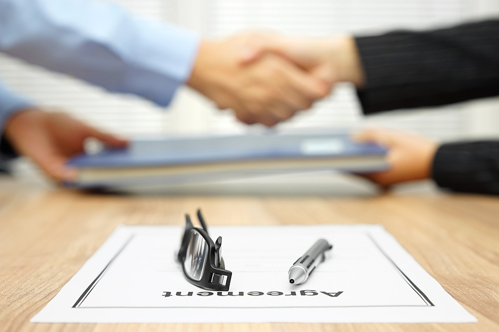Reading glass & pen sitting on agreement in front collection lawyer & client handshaking